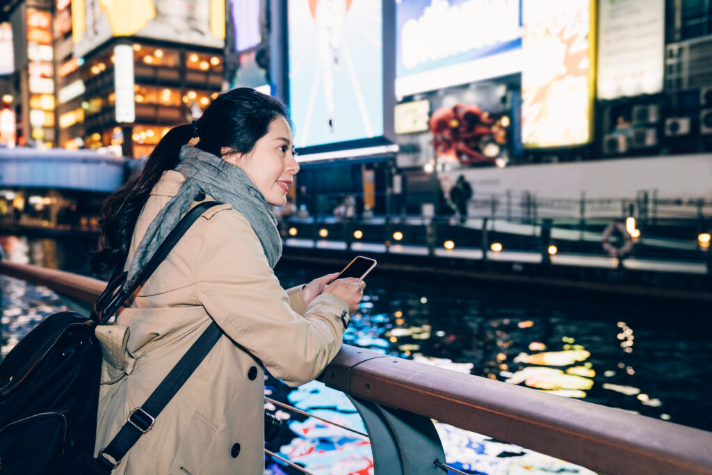 dziewczyna turysta stojący relaks nad rzeką w dotonbori osaka japonia. piękna pani podróżnik trzymając smartfon za pomocą SMS-ów online, widząc nocny widok na kanał i ruchliwe miasto.